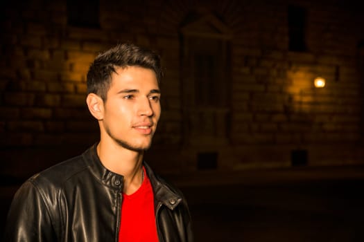 A man in a black leather jacket standing in a dark city street at night