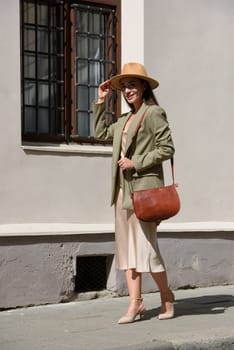 pretty brunette girl in satin long dress, hat, ankle strap shoes and checkered jacket posing with leather handbag outside.