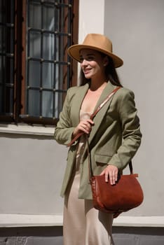 pretty brunette girl in satin long dress, hat, ankle strap shoes and checkered jacket posing with leather handbag outside.