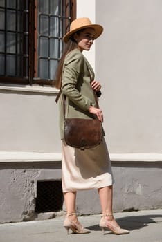 pretty brunette girl in satin long dress, hat, ankle strap shoes and checkered jacket posing with leather handbag outside.