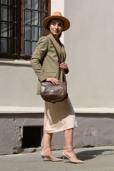 pretty brunette girl in satin long dress, hat, ankle strap shoes and checkered jacket posing with leather handbag outside.