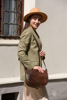 pretty brunette girl in satin long dress, hat, ankle strap shoes and checkered jacket posing with leather handbag outside.