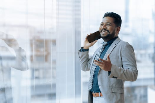 Indian businessman using smartphone talking having phone call conversation in office looking out window.