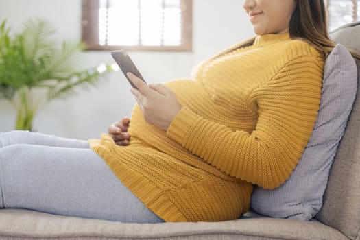 Happy Asian pregnant woman using smart phone while relaxing on sofa in her living room.