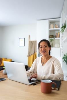 Happy Asian female entrepreneur working using laptop looking at camera. Smiling Chinese woman working with computer at home office. Vertical. Technology concept.