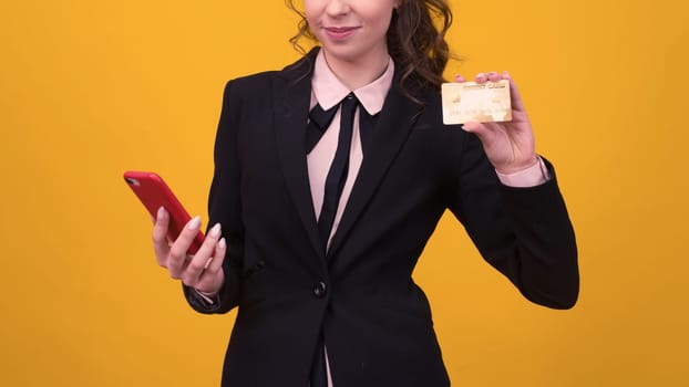 young woman posing isolated over yellow wall background using mobile phone holding debit card.