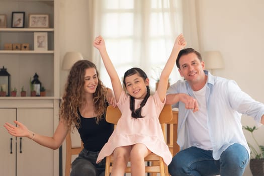 Happy family portrait with lovely little girl smiling and looking at camera, lovely and cheerful parent and their daughter sitting together in living room at home with warm daylight. Synchronos