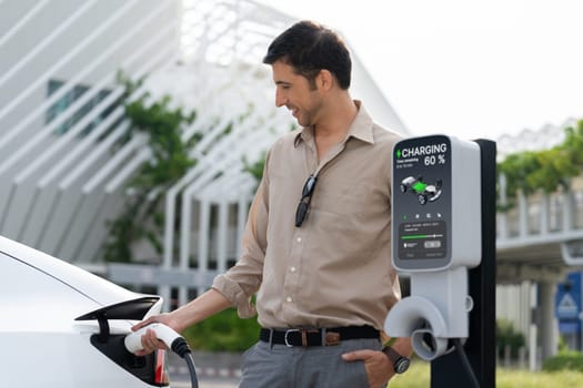 Young man put EV charger to recharge electric car's battery from charging station in city commercial parking lot. Rechargeable EV car for sustainable environmental friendly urban travel. Expedient