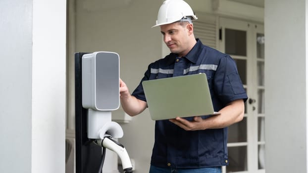 Qualified technician working on home EV charging station installation, making troubleshooting and configuration setup on charging system with laptop for EV at home. Panorama Synchronos