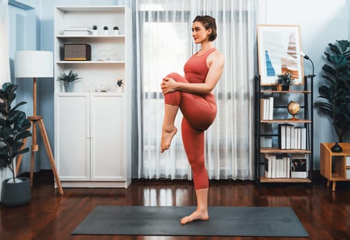 Flexible and dexterity woman in sportswear doing yoga position in meditation posture on exercising mat at home. Healthy gaiety home yoga lifestyle with peaceful mind and serenity.