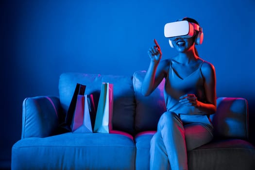 Smart female sit on sofa beside bags wear VR headset connecting metaverse, future cyberspace community technology. Elegant woman enjoy shopping products from online store in meta world. Hallucination.