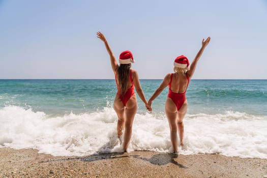 Women Santa hats ocean play. Seaside, beach daytime, enjoying beach fun. Two women in red swimsuits and Santa hats are enjoying themselves in the ocean waves and raising their hands up