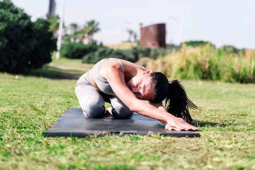 young woman in sportswear kneeling on her yoga mat doing back stretching exercises on the grass in the park, active and healthy lifestyle concept