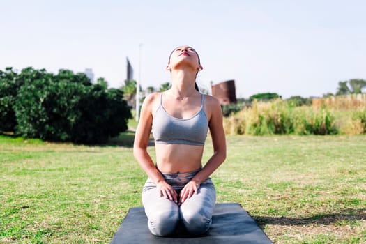 young asian woman in sportswear kneeling on her yoga mat on the park grass doing stretching and relaxation exercises, active and healthy lifestyle concept