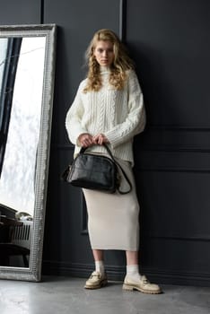 Studio portrait of beautiful woman with a curly blond hair holding black bag, posing on gray background. Model wearing stylish sweater and skirt