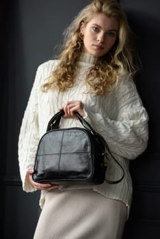 Studio portrait of beautiful woman with a curly blond hair holding black bag, posing on gray background. Model wearing stylish sweater and skirt