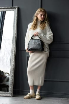 Studio portrait of beautiful woman with a curly blond hair holding black bag, posing on gray background. Model wearing stylish sweater and skirt