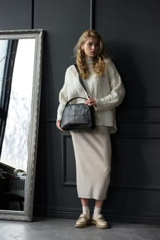 Studio portrait of beautiful woman with a curly blond hair holding black bag, posing on gray background. Model wearing stylish sweater and skirt