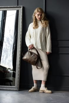 Studio portrait of beautiful woman with a curly blond hair holding brown bag, posing on gray background. Model wearing stylish sweater and skirt