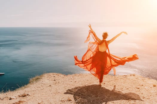 Side view a Young beautiful sensual woman in a red long dress posing on a rock high above the sea during sunrise. Girl on the nature on blue sky background. Fashion photo.