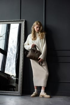 Studio portrait of beautiful woman with a curly blond hair holding brown bag, posing on gray background. Model wearing stylish sweater and skirt