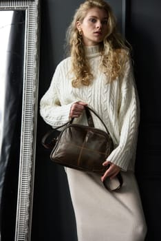 Studio portrait of beautiful woman with a curly blond hair holding brown bag, posing on gray background. Model wearing stylish sweater and skirt