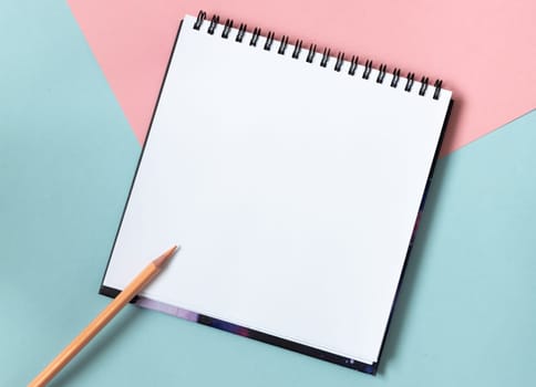 Mock up of empty spiral sketchbook with white paper on pastel pink and blue background. Top view of open notebook with clean sheet and pencil. Template for message. Copy space