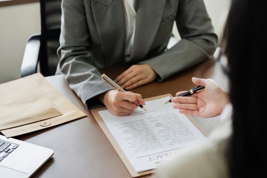 A woman is signing a document at a desk with a laptop and a chair. The woman is wearing a suit and is writing with a pen. The scene suggests a professional setting