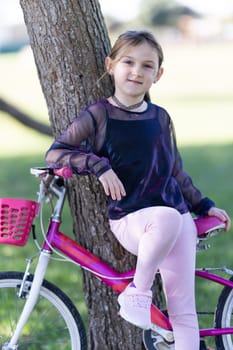 A young girl is sitting on a pink bicycle with a basket. She is wearing a black shirt and pink pants