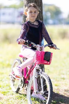 A young girl is sitting on a pink bicycle with a basket. She is wearing a black shirt and pink pants