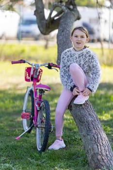 A girl is sitting on a tree branch with a pink bicycle behind her. She is wearing a white sweater and pink pants