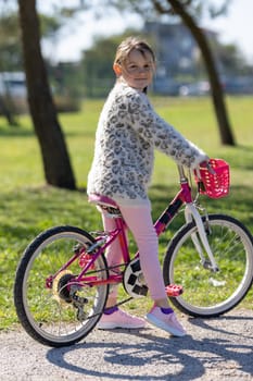 A young girl is sitting on a pink bicycle with a basket. She is wearing a white sweater and pink pants