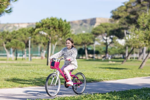 A young girl is riding a pink bike in a park. The park is full of trees and grass, and there are a few benches scattered around. The girl is wearing a pink jacket and pink pants