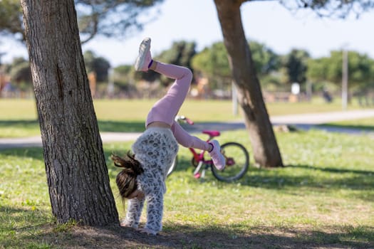 A girl is doing a handstand on a tree trunk in a park. A bicycle is parked nearby