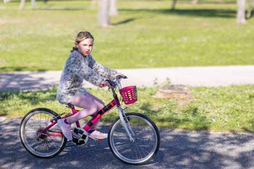 A young girl is riding a pink bicycle with a basket on the front. She is wearing a pink sweater and pink pants