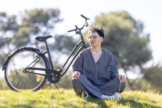 A woman is sitting on the grass next to a bicycle. He is wearing a grey shirt and grey pants