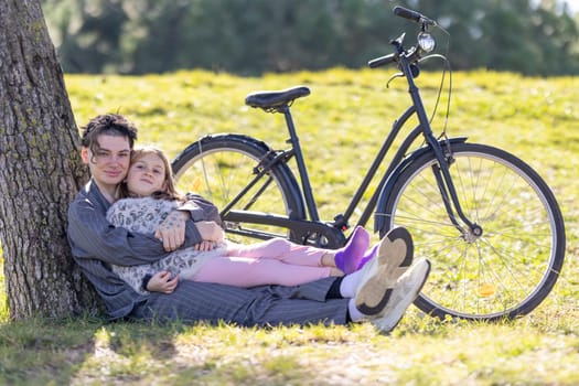 A mother and a little girl are sitting on the grass next to a bicycle. The man is holding the girl and they both seem to be enjoying the moment