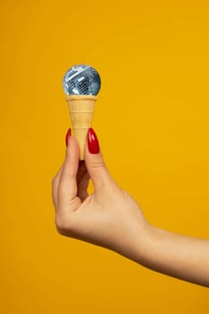 Woman hand holding small crispy ice cream cone with a mirrored disco ball isolated on yellow background