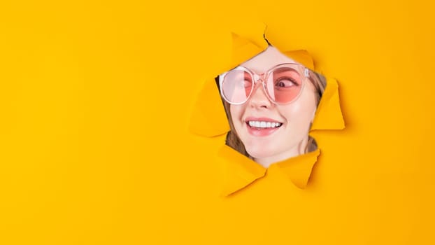 Beautiful face of young smiling woman with perfect teeth looking in paper torn hole in yellow background with copy space.