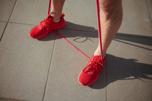 View from above athletic man in red sports shoes, doing physical exercises with red resistance band. Active healthy lifestyle. Feet in red sneakers working out with fitness band. Sports equipment