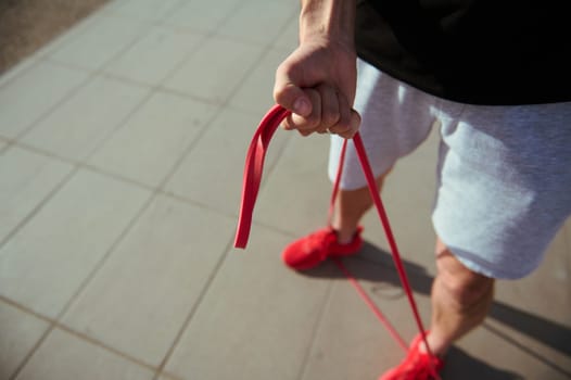Top view of a sportsman in red sneakers, gray sports shorts and black t-shirt doing warm up exercises with resistance band. Athletic man exercising outdoor with fitness band. Sport and people concept