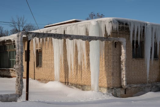 Big icicles on the roof of a townhouse on a snowy winter day among thaw. Cleaning the roofing from snow and icicles.