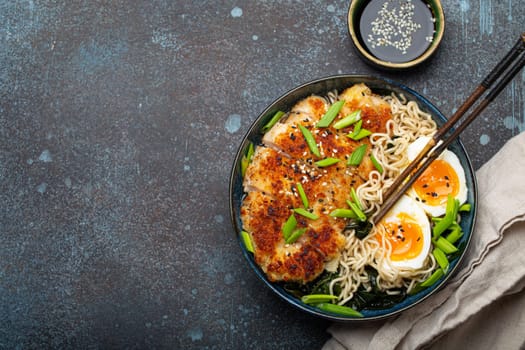 Asian noodles ramen soup with deep fried panko chicken fillet and boiled eggs in ceramic bowl with chop sticks and soy sauce on stone rustic background top view, space for text