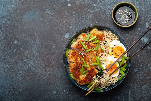 Asian noodles ramen soup with deep fried panko chicken fillet and boiled eggs in ceramic bowl with chop sticks and soy sauce on stone rustic background top view, space for text