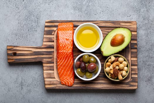 Food sources of healthy unsaturated fat: fresh raw salmon fillet, avocado, olives, nuts on cutting board on rustic stone background top view. Omega 3 and oil products, healthy nutrition and keto diet