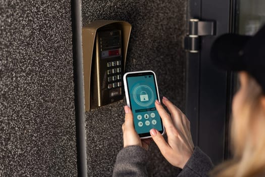 Female hand holding black mobile smart phone with smart home application on the screen. Blurred house on the background. For access to all of the controls of your house and caring of home security. High quality photo