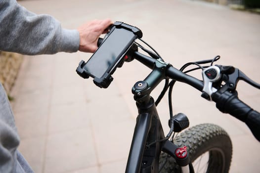 Cyclist sets up adjustments on smartphone, standing near electric bike, before riding in city. Man using mobile phone, renting electric bike using rental app. Bike sharing city service. Mockup screen