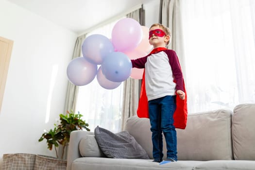Boy in superhero costume with red cape and mask holding balloons. Home interior casual portrait. Childhood imagination and play concept. Design for greeting card, poster, invitation