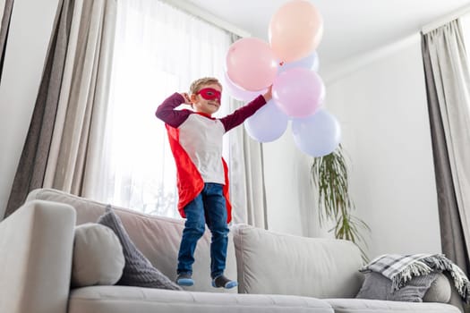 A young child dressed as a superhero stands proudly on a sofa, joyfully playing with colorful balloons.