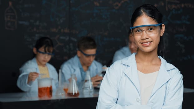 Girl looking at camera with arm folded while people doing experiment at laboratory. Cute student standing blackboard with chemical theory with blurring background at STEM science class. Edification.